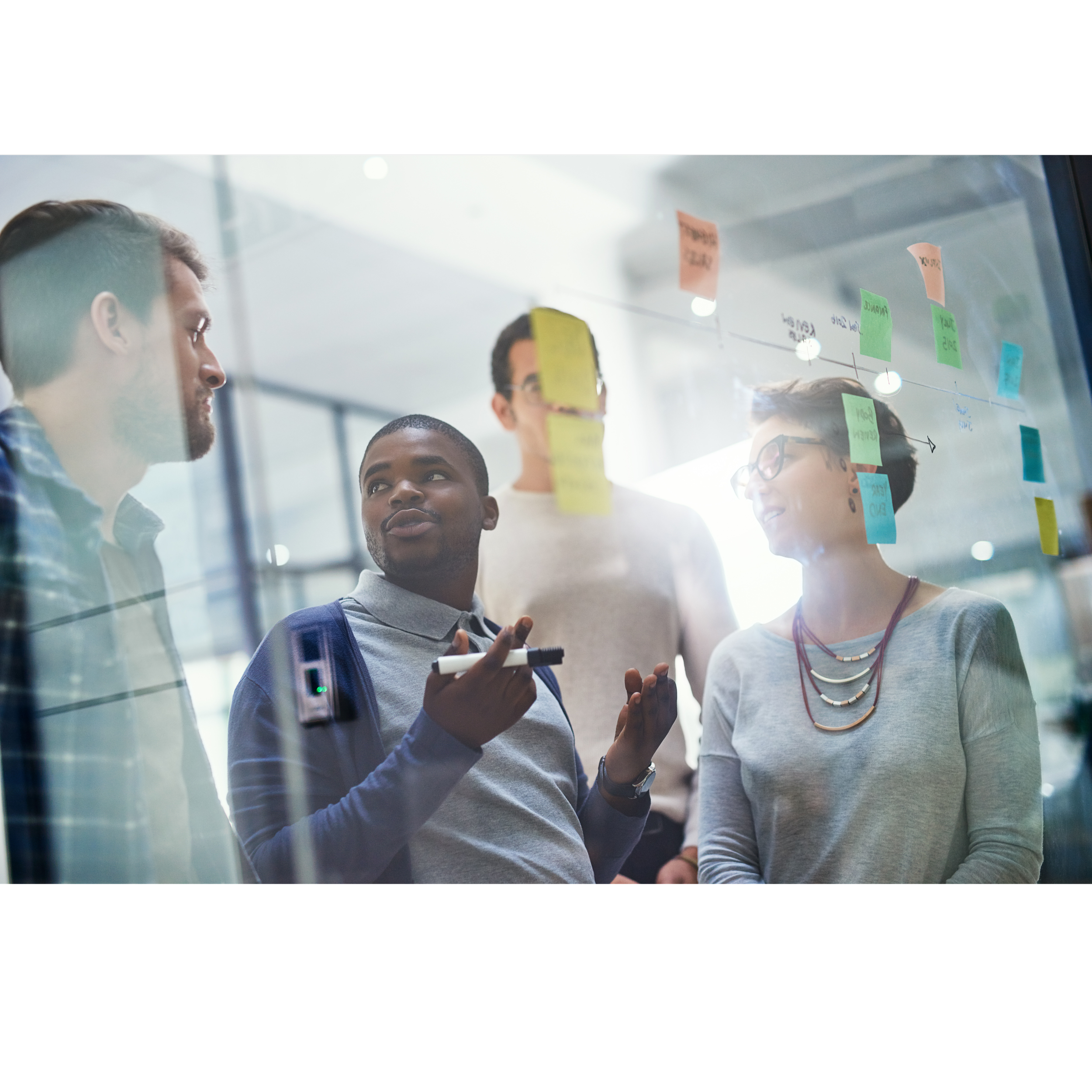 Four diverse professionals collaborating in a modern office, facing a glass wall covered with colorful sticky notes and diagrams.