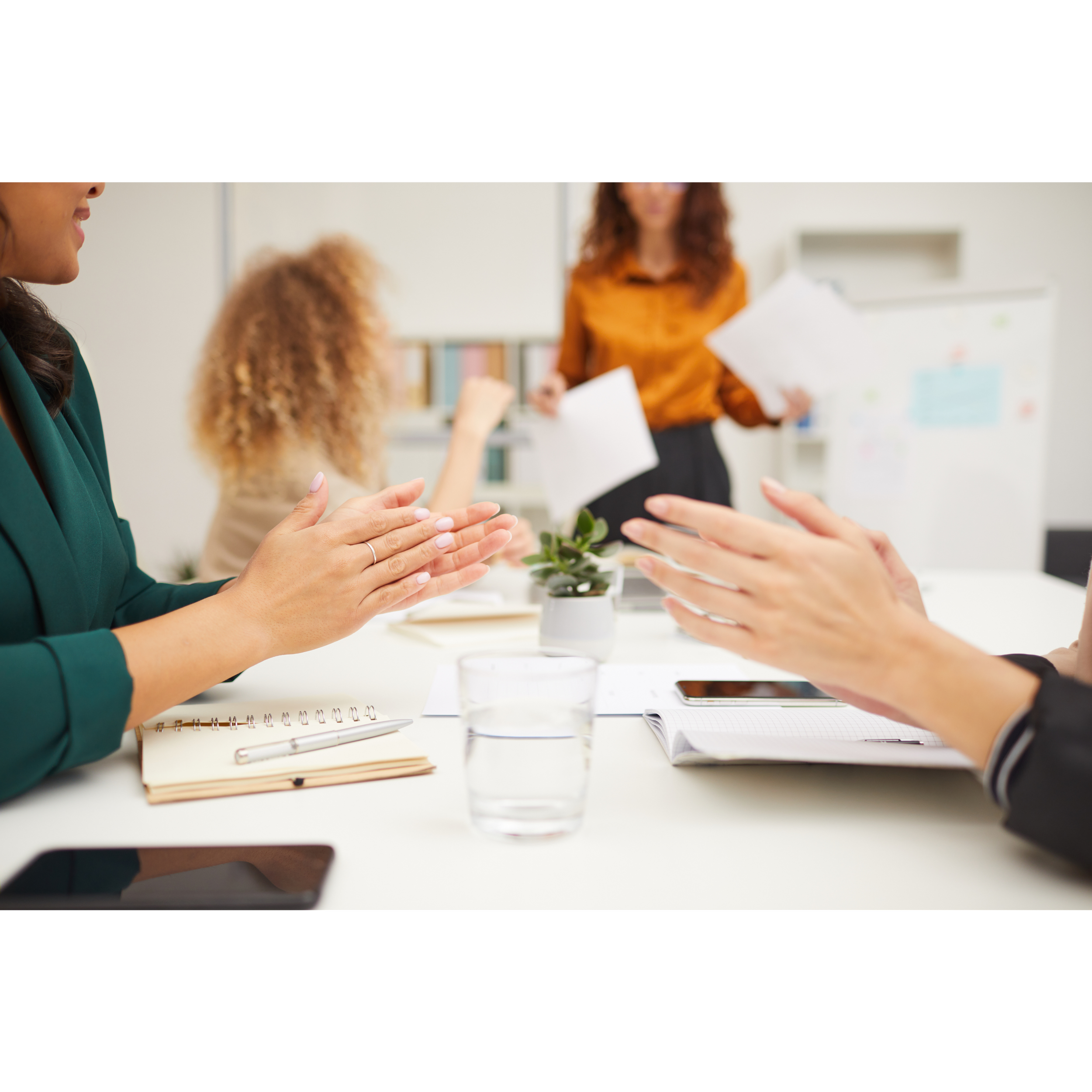People in a meeting clapping while a woman in an orange shirt speaks in the background.