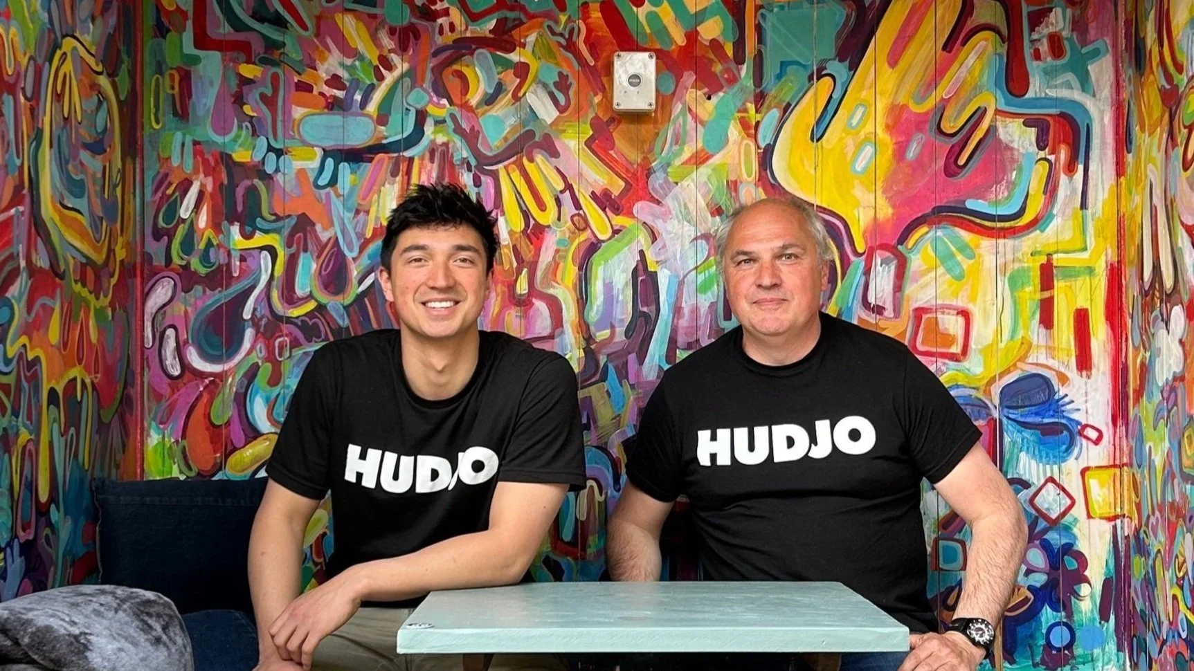 Two men sitting at a table in front of a colorful abstract wall. Both are wearing black shirts with the word 'HUDJO' printed on them.