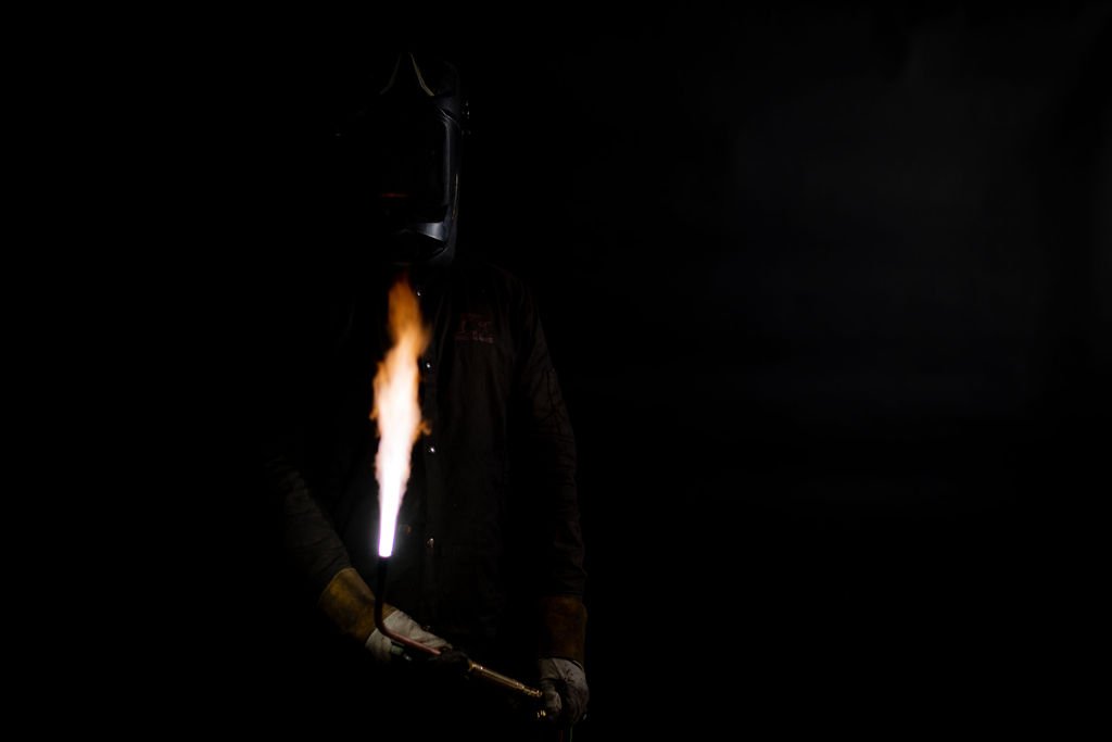 A helmeted welder holding a lit torch. He is dressed in all black and in front of a black backdrop