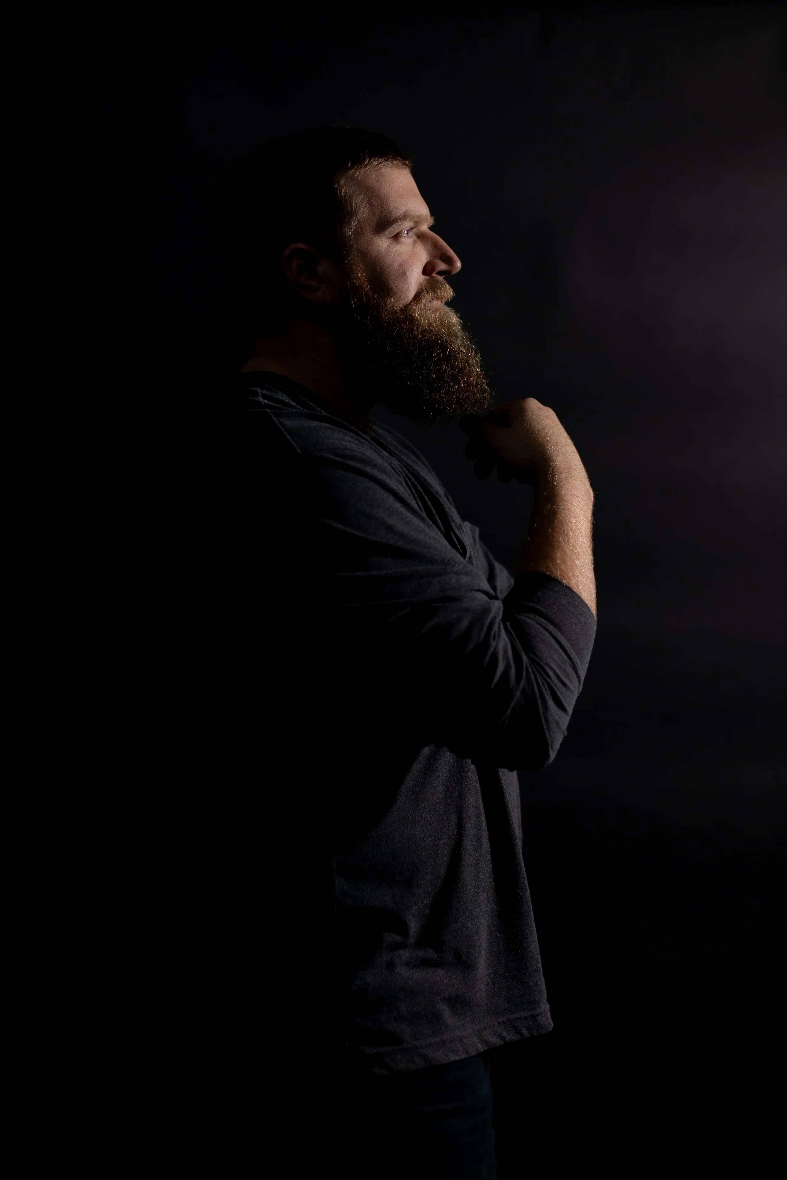Headshot of a bearded man touching his beard. The backdrop is black.