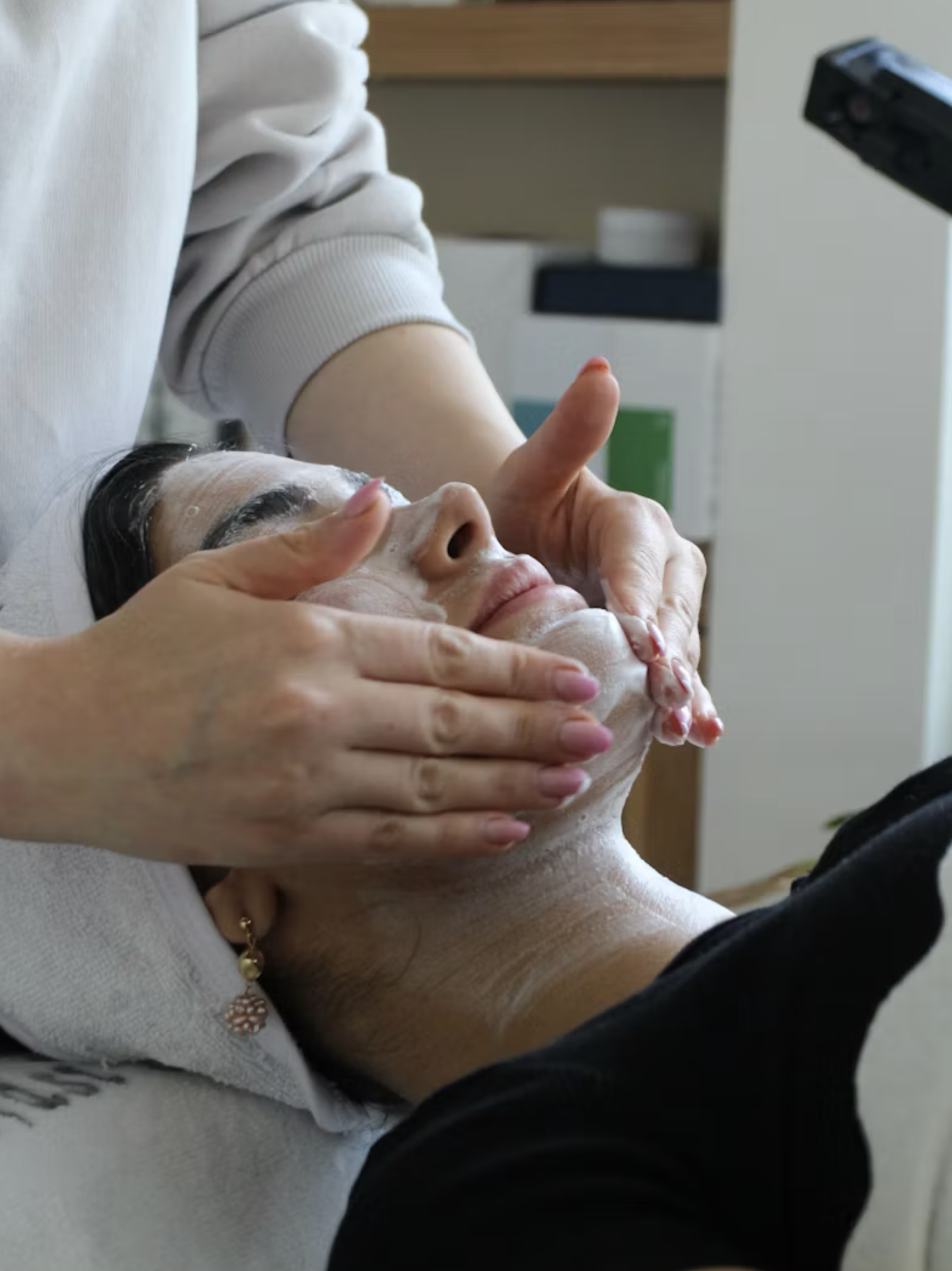 A person is receiving a facial treatment with soap or foam applied to their face by a professional, in a spa or clinic setting.