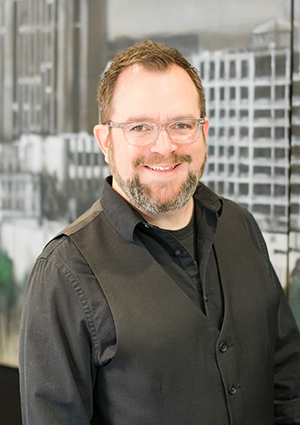 A man with glasses, a beard, and short hair smiling, wearing a dark vest over a black shirt, standing in front of an office window with a cityscape view.