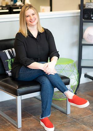 A woman with blonde hair sitting on a black bench, smiling. She is wearing a black shirt, blue jeans, and red shoes. There is a green side table and various objects in the background.