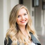 Portrait of a young woman with long blonde hair, smiling and wearing a black blazer, standing outside near a building