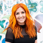 A woman with long, wavy red hair smiling in front of a colorful, artistic background.