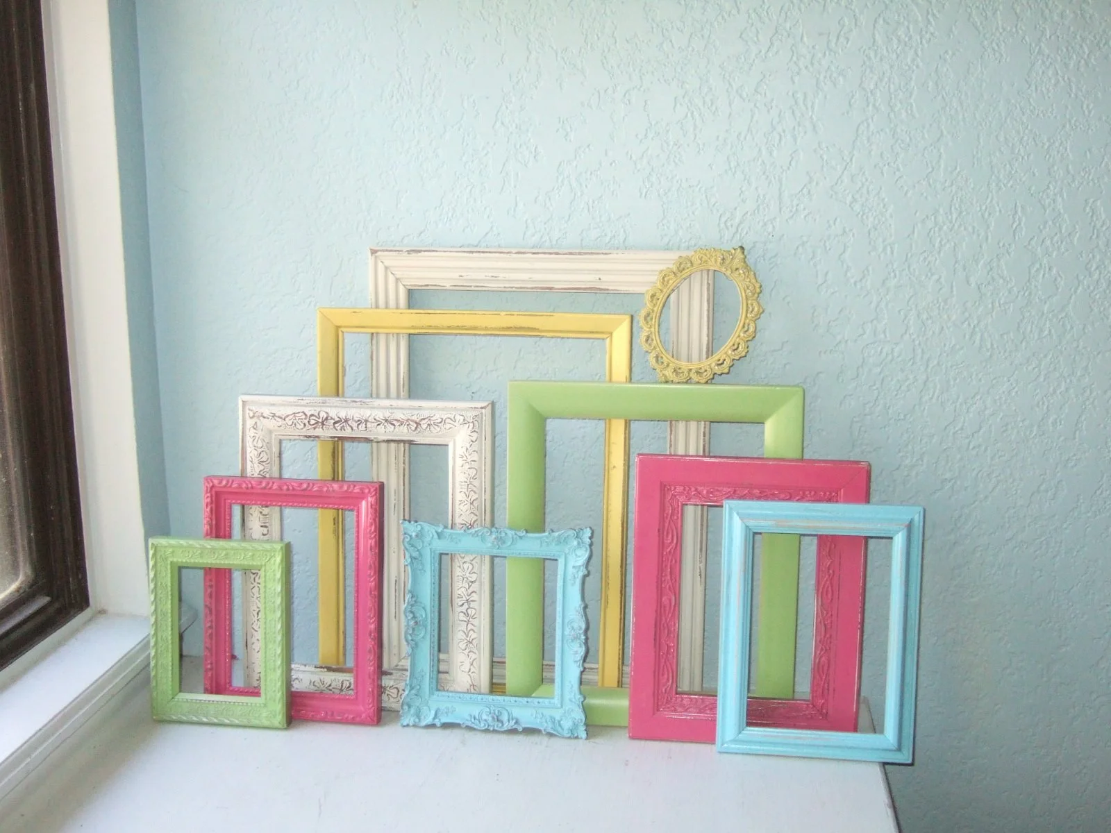 A collection of empty colorful picture frames arranged in front of a textured blue wall on a white surface, near a window.