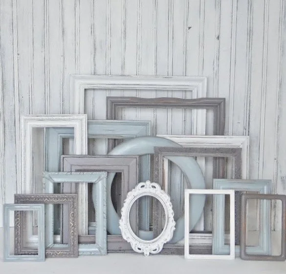 A collection of empty picture frames of various sizes and colors arranged against a white wooden wall.