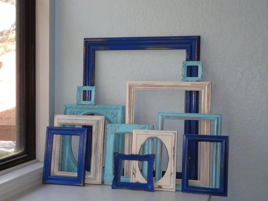 Multiple empty picture frames in blue, white, and beige colors are arranged on a white surface near a window with a light-colored wall behind them.