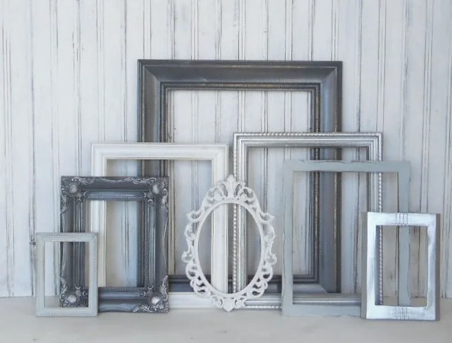 Multiple empty picture frames of various sizes and styles leaning against a white wooden wall.