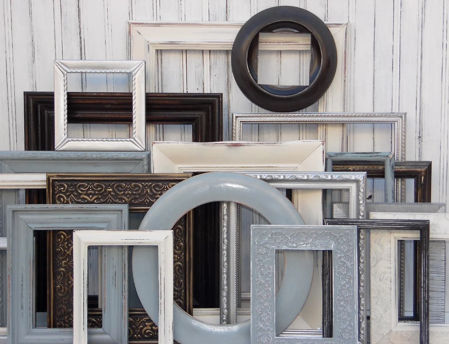 Various empty picture frames of different shapes, sizes, and colors leaning against a white wooden wall.