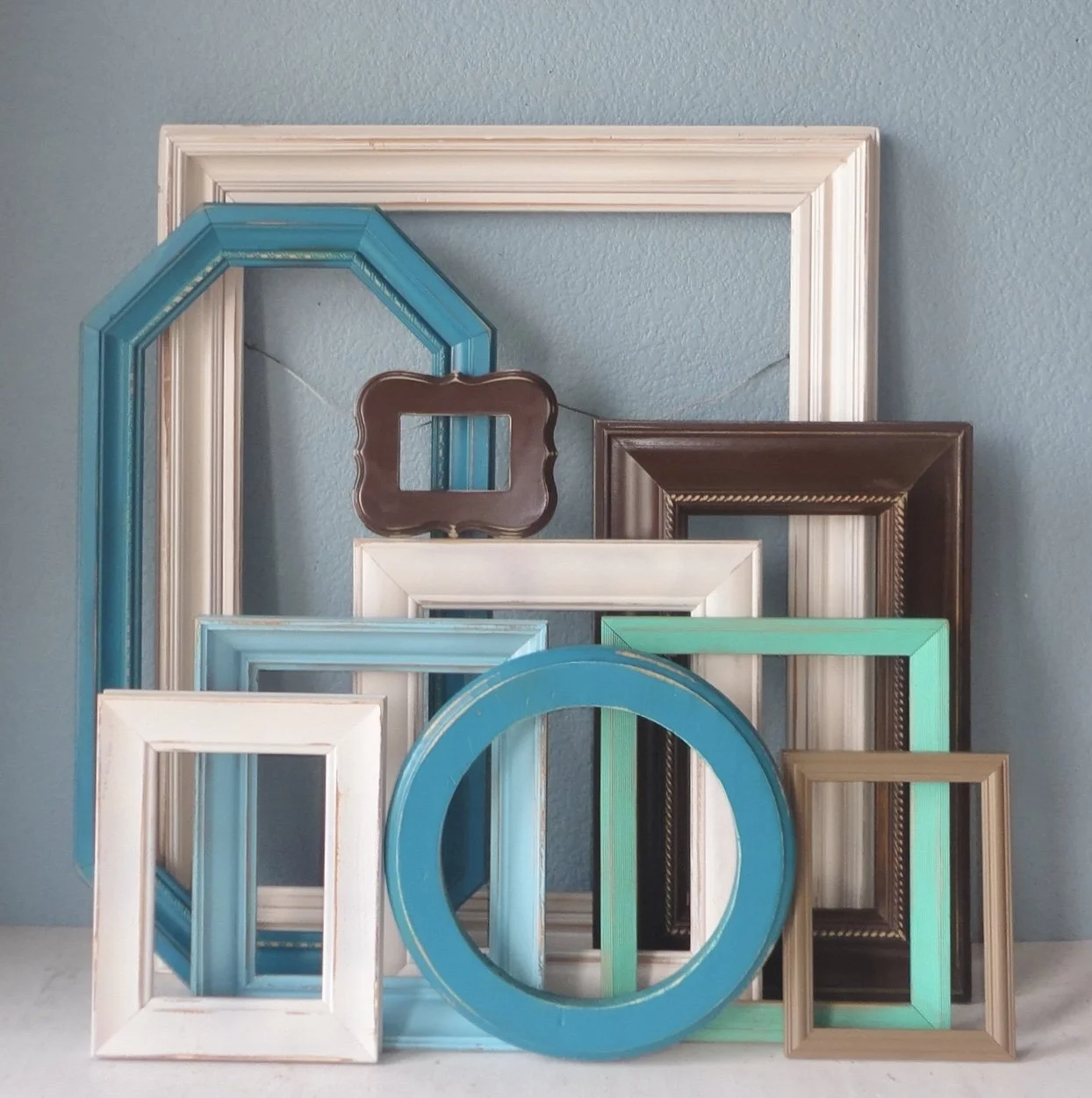 Multiple empty picture frames of various sizes and colors arranged against a blue wall, some leaning and some hanging, including white, brown, teal, and gold frames.