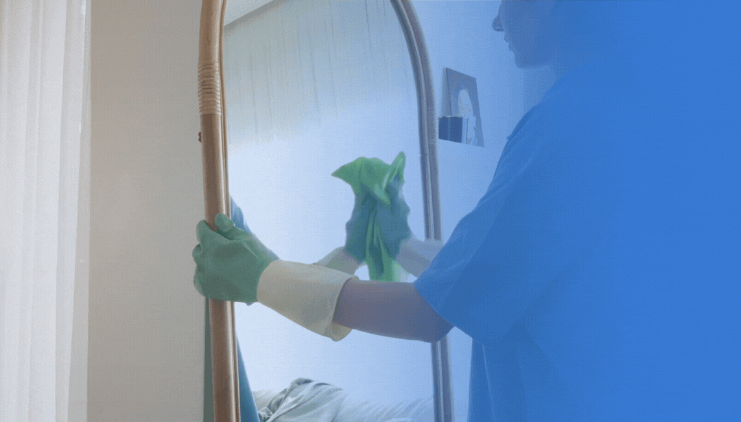 A person wearing gloves and scrubbing a mirror with a green cloth in a bedroom.