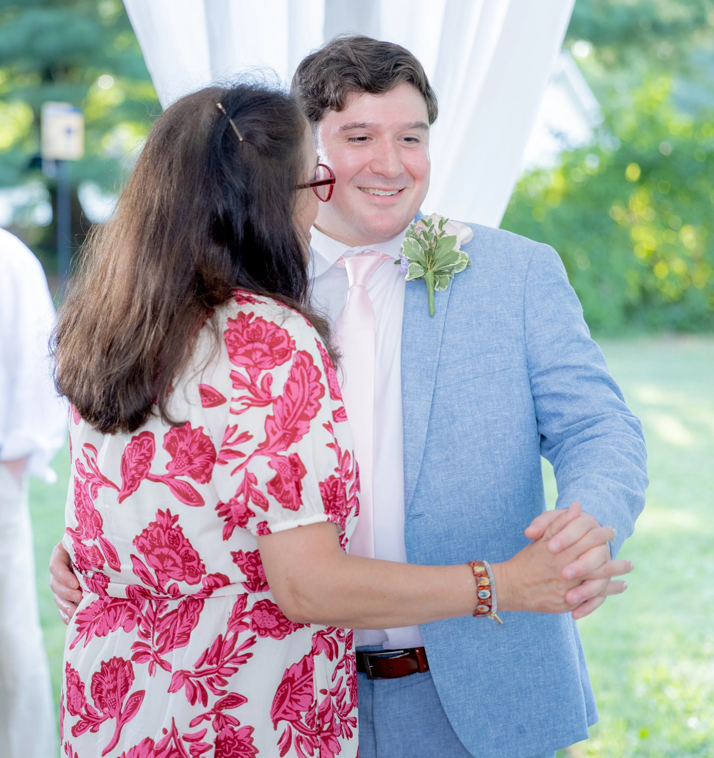 A man and woman holding hands and smiling at each other outdoors, with white drapes and green trees in the background