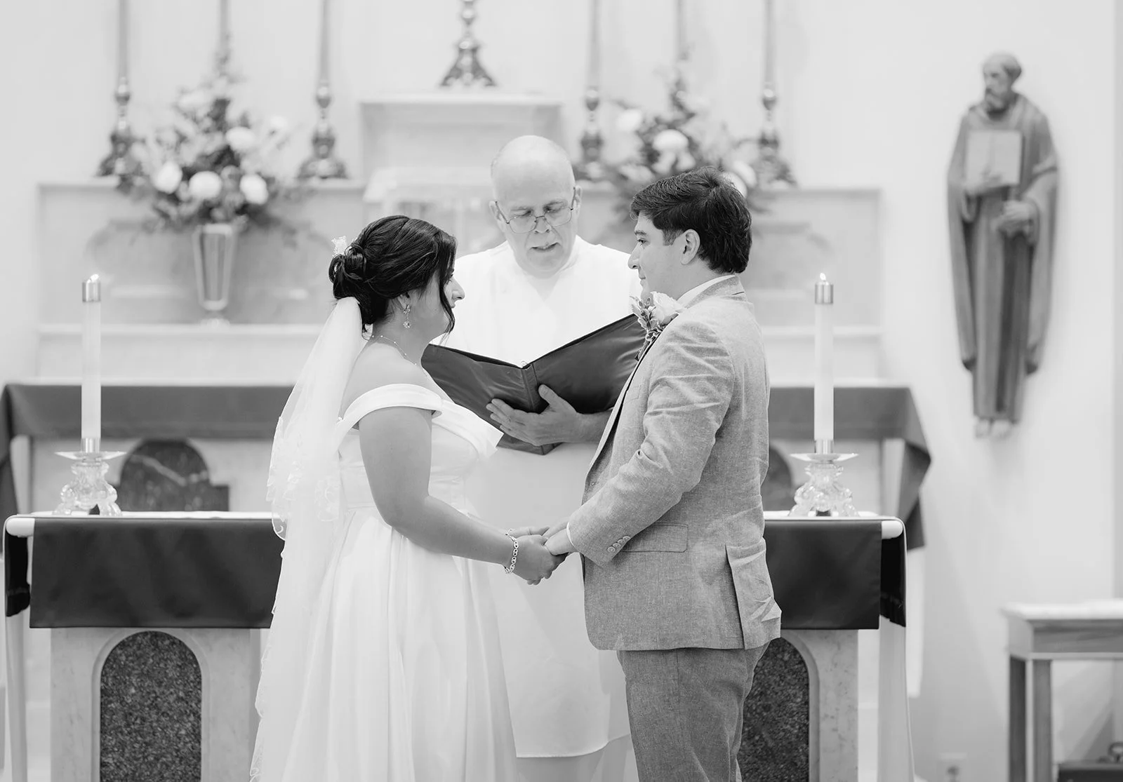A black and white photo of a couple holding hands during a wedding ceremony in a church, with an officiant reading from a book and religious statues and candles in the background.