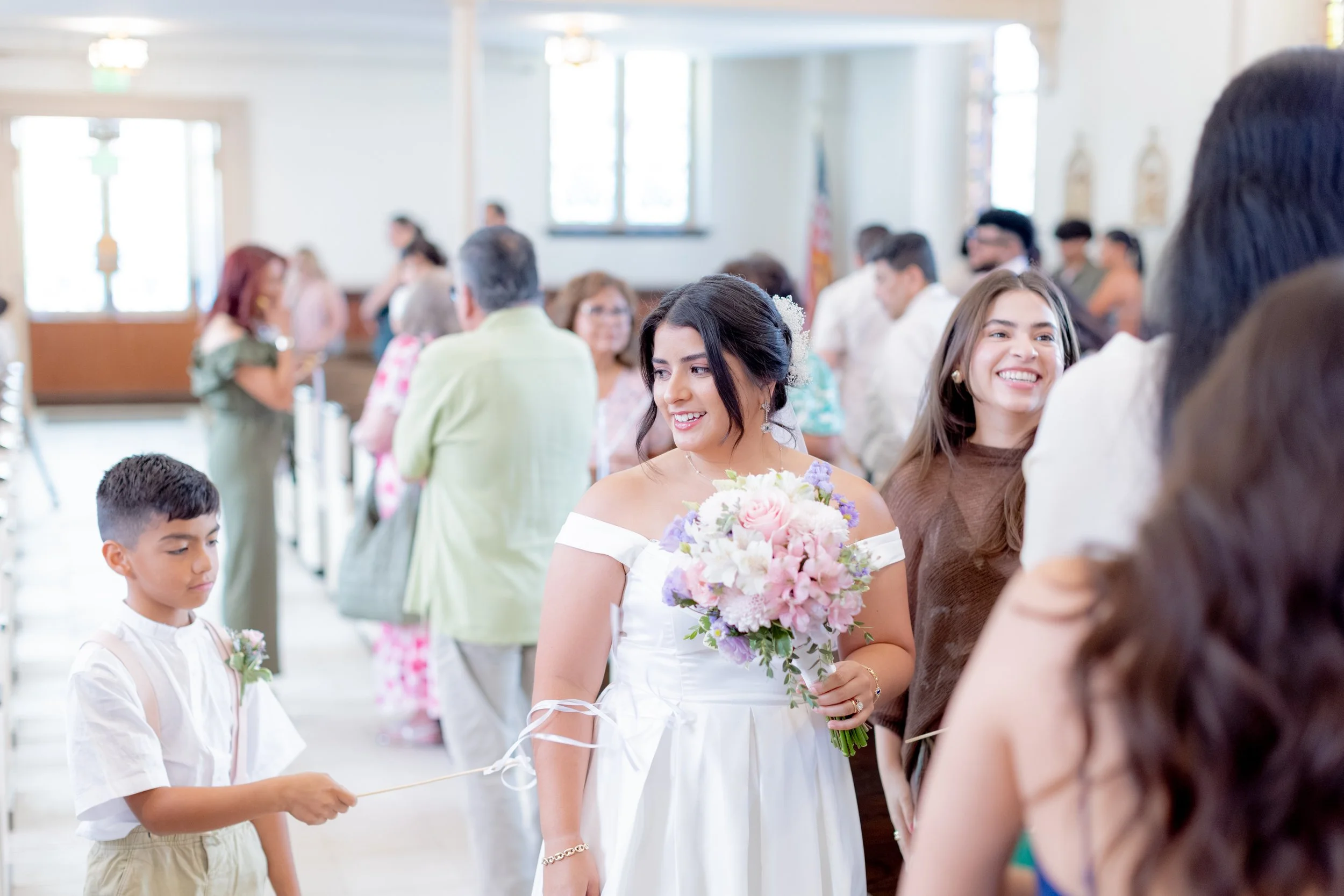 A bride holding a bouquet of pink and purple flowers, smiling and talking to a man in a dark suit at her wedding reception. The reception hall is filled with guests of various ages and ethnicities, some holding small bouquets or wearing boutonnieres, with large windows and bright lighting.