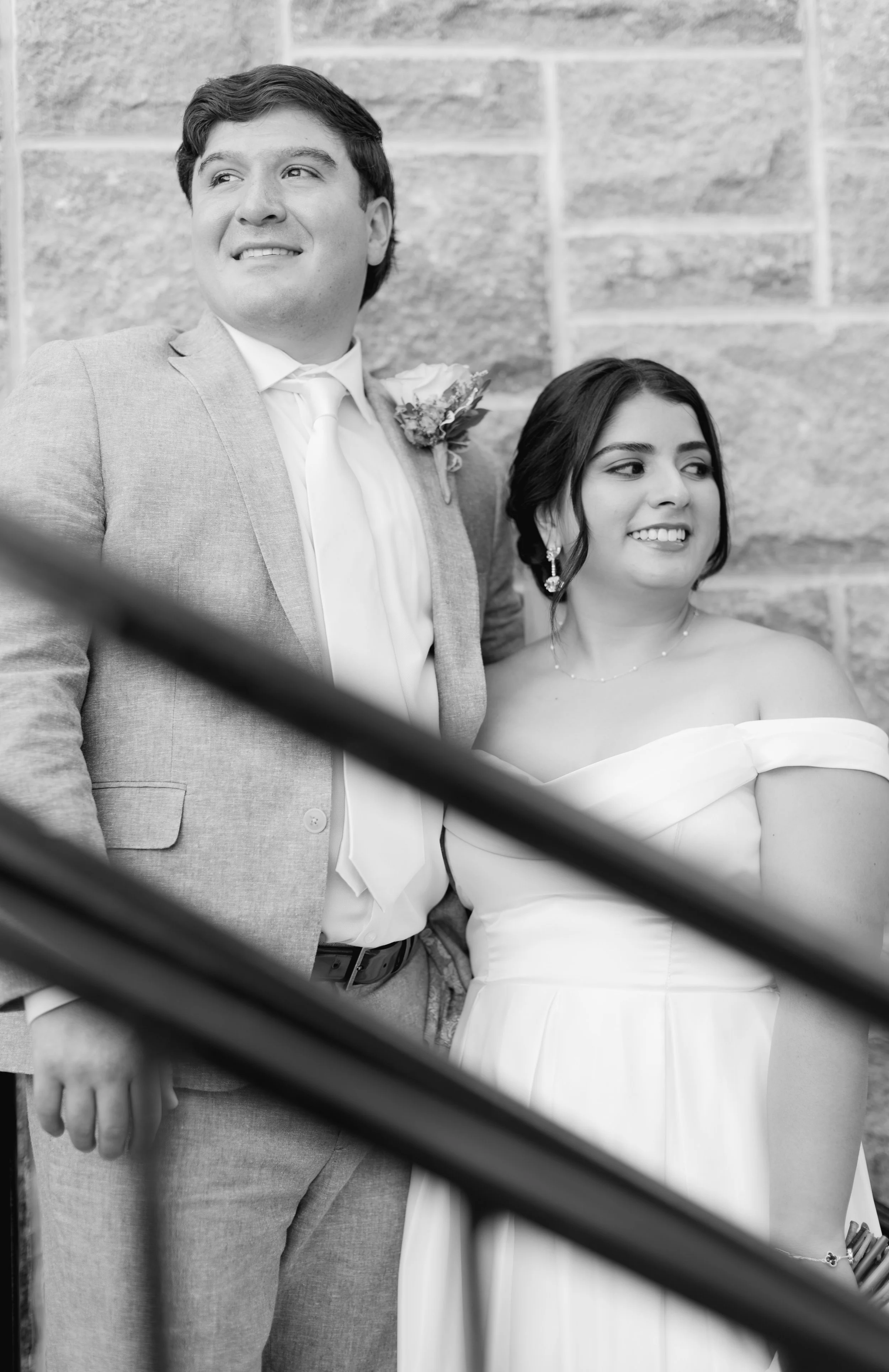 Black and white photo of a smiling young man in a light-colored suit and a woman in an off-shoulder dress standing outdoors against a brick wall, partially obscured by railing.