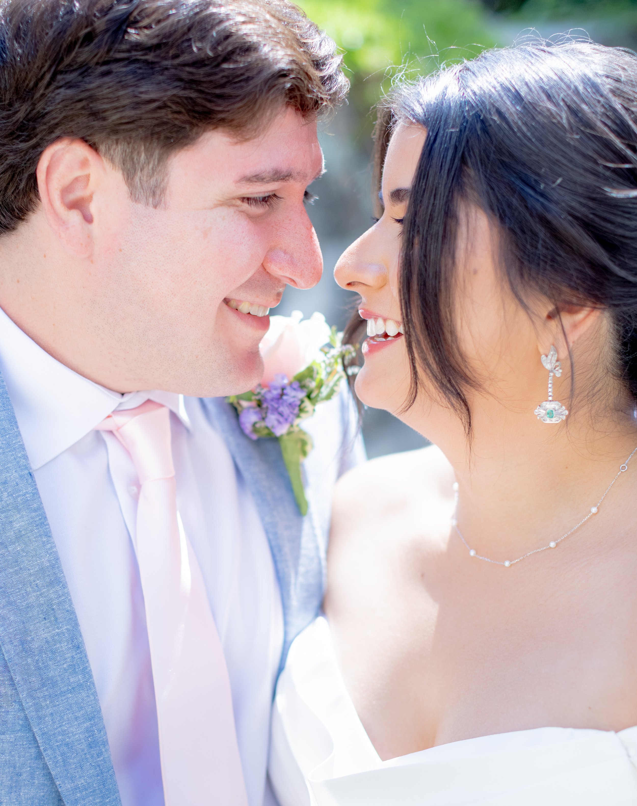 A close-up of a smiling couple with foreheads touching, outdoors with greenery in the background, dressed in wedding attire.