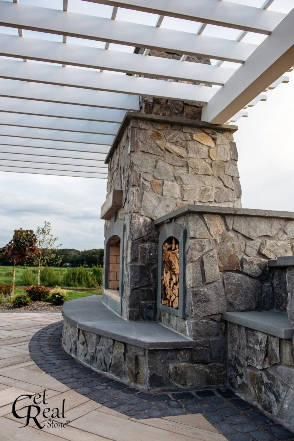 A stone outdoor fireplace and wood storage under a white pergola, with a landscape and trees in the background.
