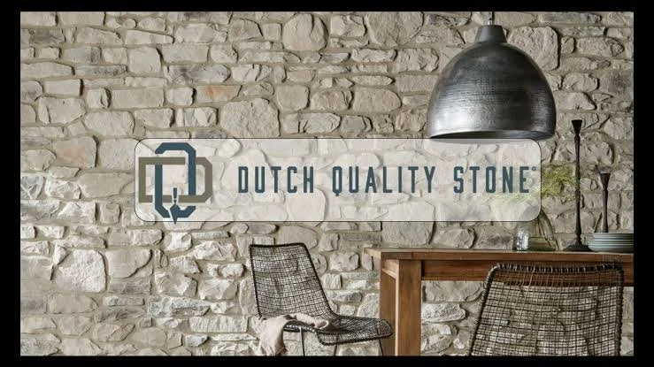 Interior of a room with a stone wall, a wooden table, metal chair, and large metallic pendant light, with a sign reading 'Dutch Quality Stone' on the wall.
