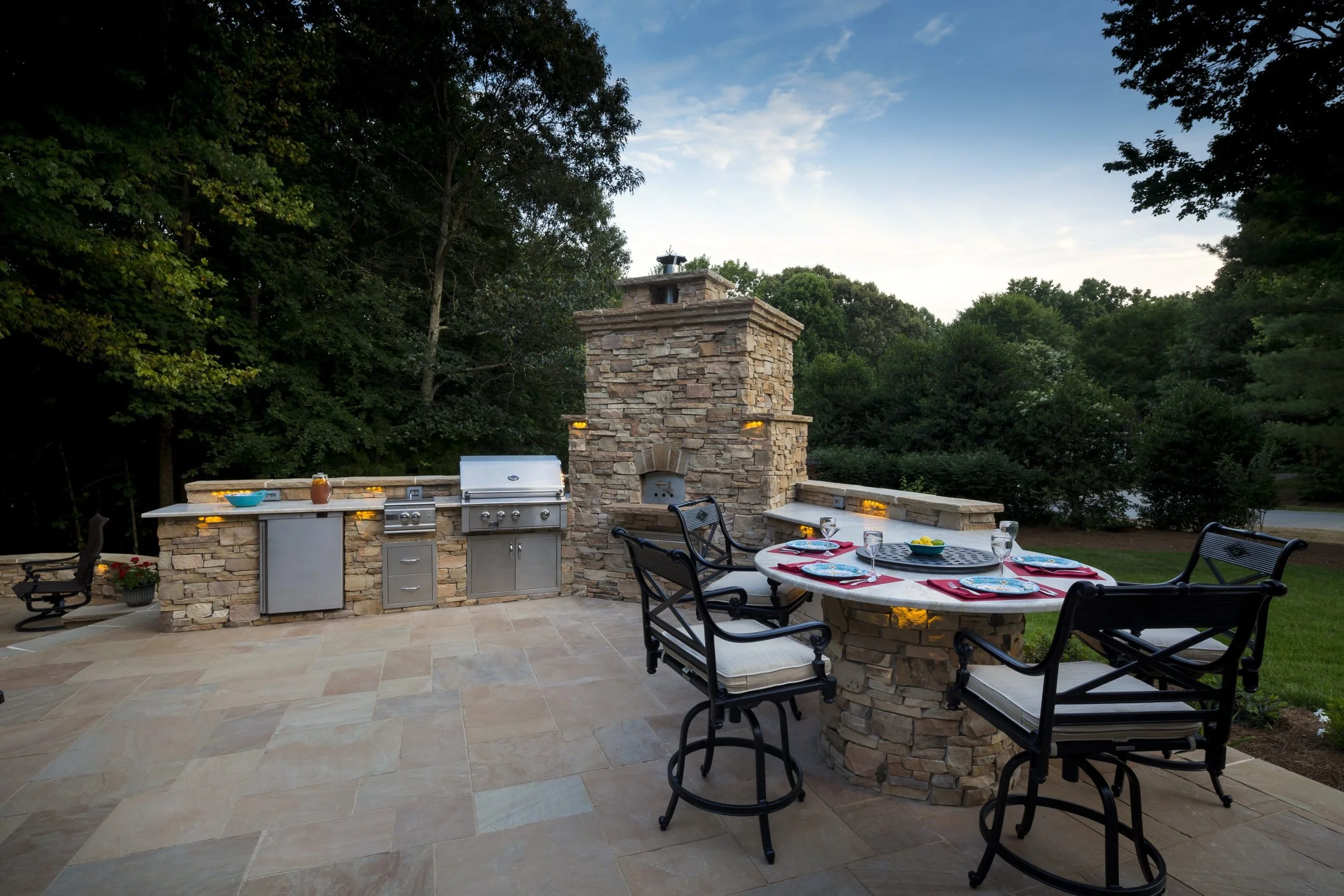 An outdoor patio with a stone kitchen counter, grill, and chimney, surrounded by trees, with a table set for four with plates, napkins, and glasses, and a scenic view of the sky and forest.