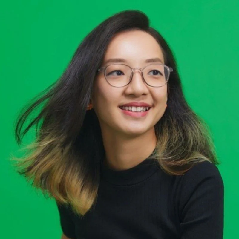 Young woman with glasses and long hair smiling in front of a green screen.