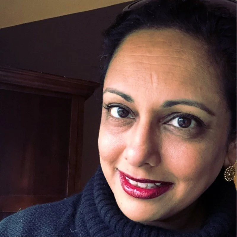 A woman with dark hair, wearing a dark turtleneck sweater, smiling, with bright lipstick, earrings, and makeup.