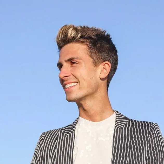 A young man with styled blond and brown hair, smiling, wearing a black-and-white striped blazer and a white shirt, against a clear blue sky background.