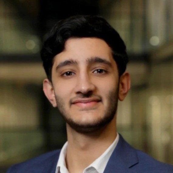 A young man with dark hair and a trimmed beard, wearing a navy blazer and white shirt, standing in front of a blurred professional or office environment.