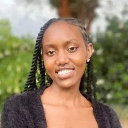 A young woman with twists in her hair smiling outdoors with greenery and trees in the background.