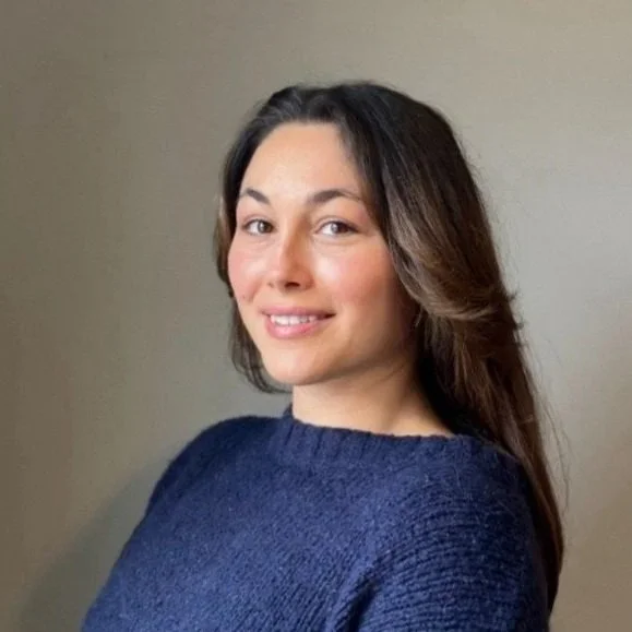 A young woman with long brown hair and a smile, wearing a navy blue sweater, standing against a plain beige background.