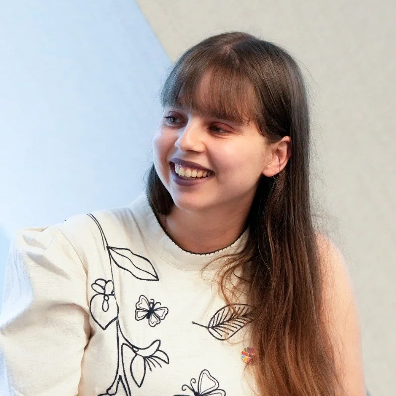 Young woman with long brown hair and bangs smiling, wearing a white top with leaf and butterfly illustrations.