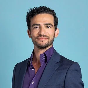 Headshot of a man with dark curly hair and a beard, wearing a dark blue suit jacket and a purple shirt, smiling against a light blue background.