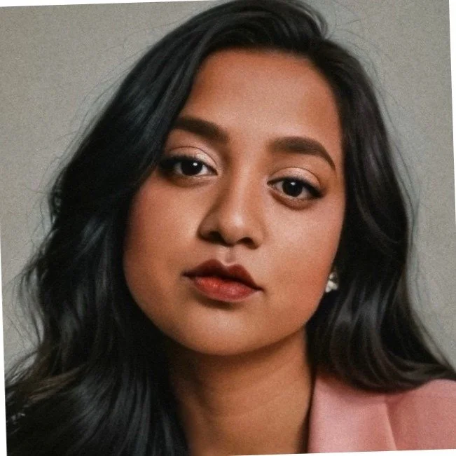 A close-up portrait of a woman with long black hair, light makeup, and a pink blazer, looking at the camera.