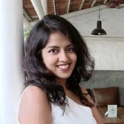 A smiling woman with dark wavy hair wearing a beige sleeveless top indoors with a wooden ceiling and hanging light fixture.