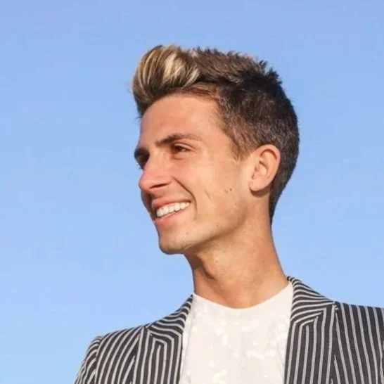 A young man with styled blond and brown hair, smiling, wearing a black-and-white striped blazer and a white shirt, against a clear blue sky background.