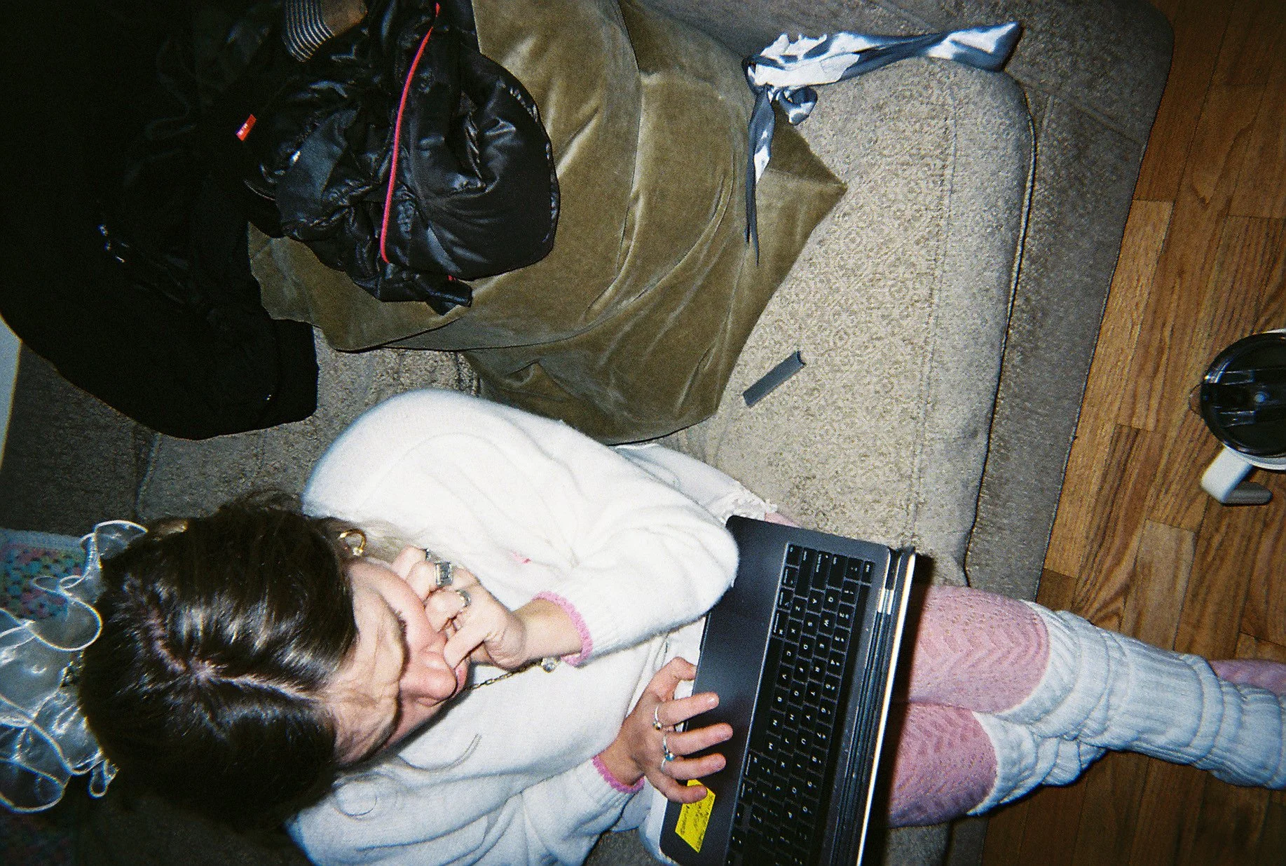 A woman with dark hair styled with a bow, wearing a white fuzzy sweater and pink patterned leg warmers, kneeling on a wooden floor, using a black laptop. Behind her is a couch with several bags and clothing items on it.