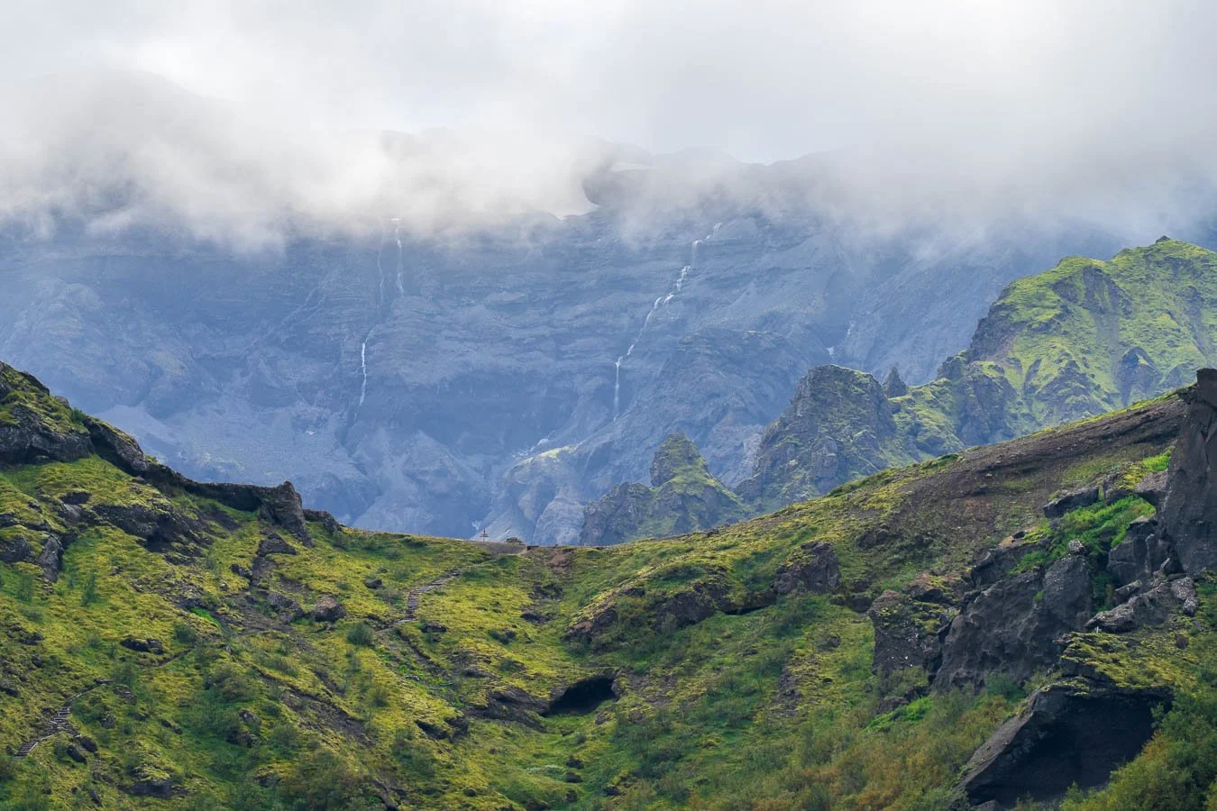 Telephoto view in Þórsmörk showing green ridges beneath misty glacier-carved cliffs, with multiple thin meltwater waterfalls descending from low cloud near the Fimmvörðuháls trail.