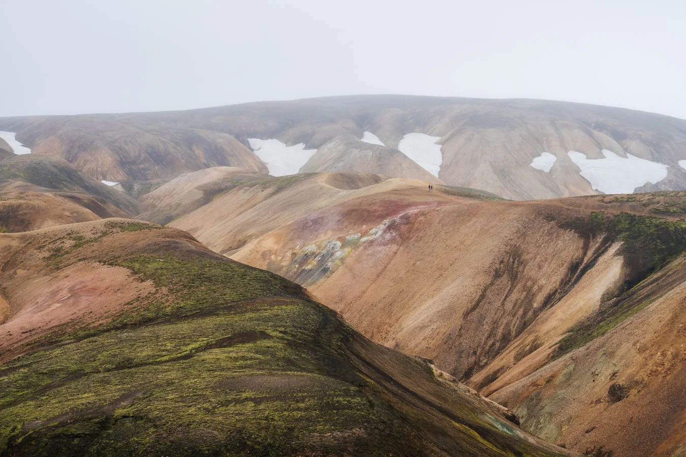 Moody scene of tiny hikers on the Laugavegur trail between Hrafntinnusker and Landmannalaugar, walking through expansive rhyolite hills in dense fog and overcast conditions in Iceland’s Highlands.