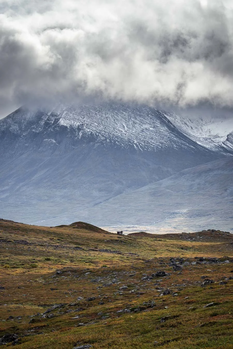 Telephoto view of a small alpine hut along the Kungsleden Trail in Swedish Lapland, set against a dramatic mountain backdrop at higher elevation.