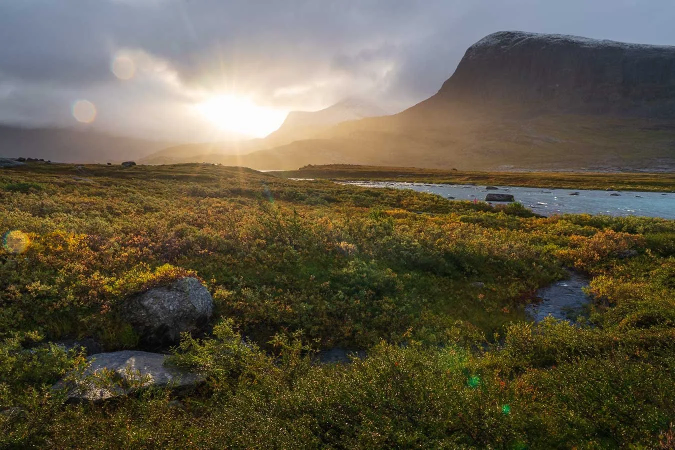 Sunrise along the Kungsleden trail between Abiskojaure and Alesjaure in Swedish Lapland, featuring a sunburst emerging through partial cloud cover beside rugged mountain peaks in the distance.