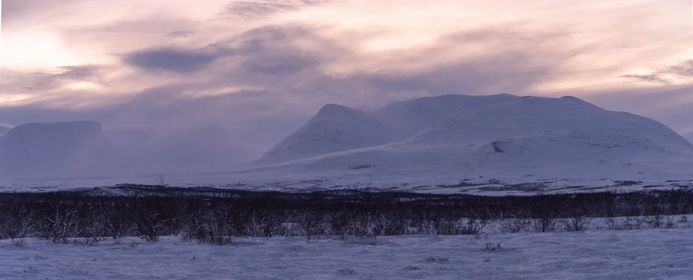 Dramatic sunrise view in Abisko National Park, Northern Sweden, showing distant mountains partially obscured by dense clouds with sunlight breaking through, creating striking lighting and atmospheric contrast.