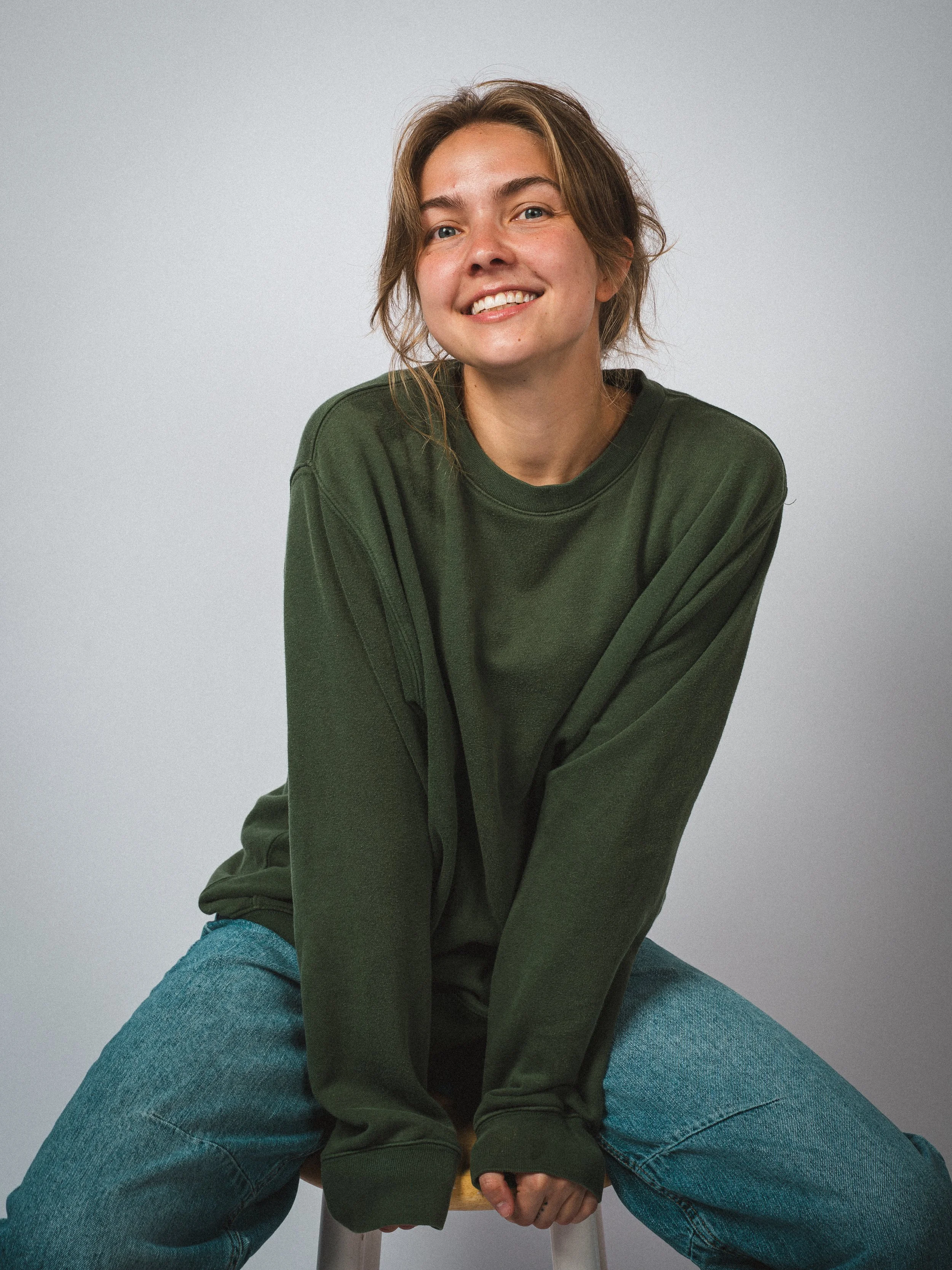 Portrait of a smiling young woman sitting on a stool with legs spread, wearing a dark green sweatshirt and blue jeans, against a plain light gray background.
