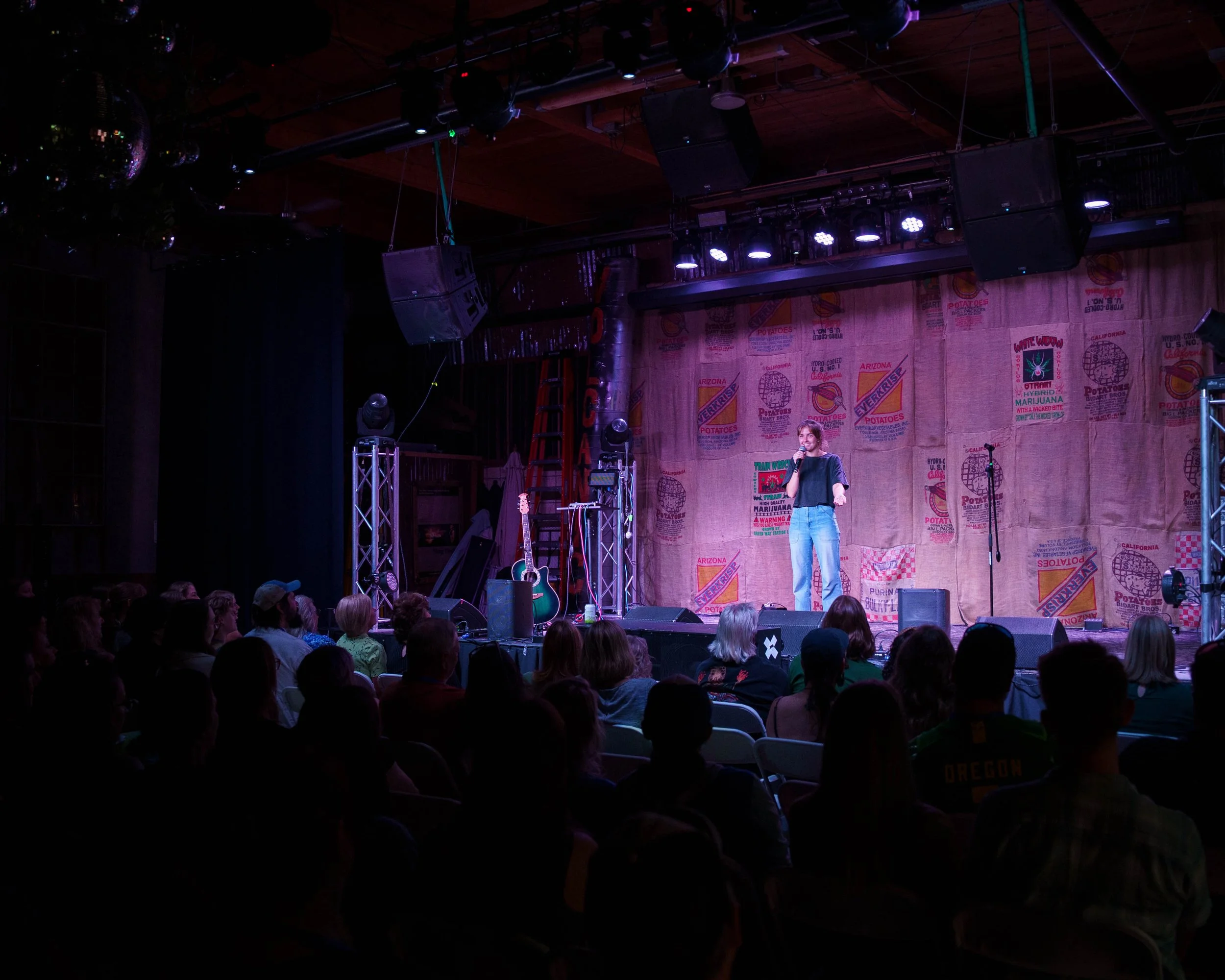 A woman standing on stage holding a microphone in a dimly lit venue with audience seated in front, stage equipment, and a wall with colorful posters in the background.