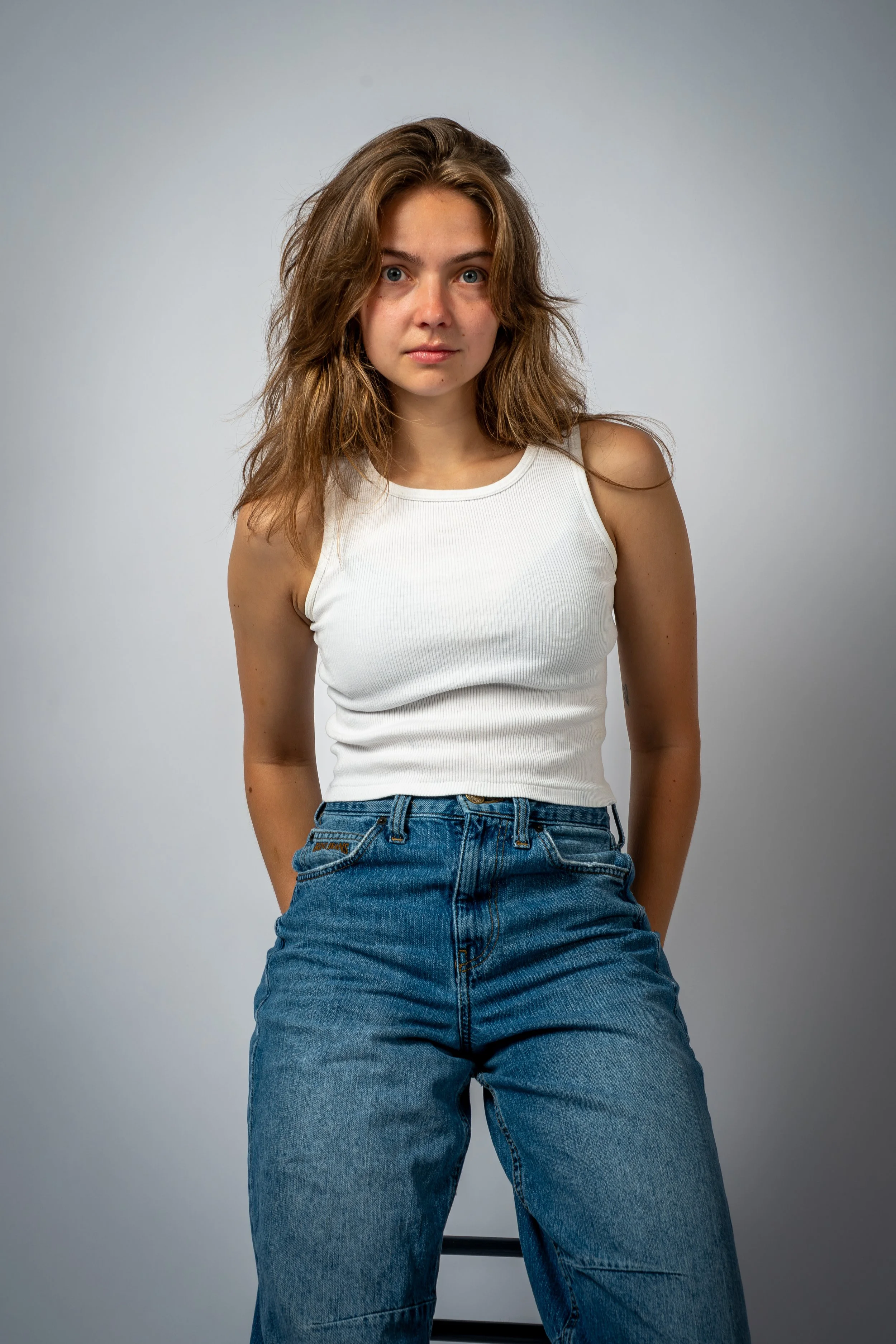 A young woman with wavy brown hair and blue eyes, wearing a white sleeveless top and blue jeans, standing against a plain light gray background.
