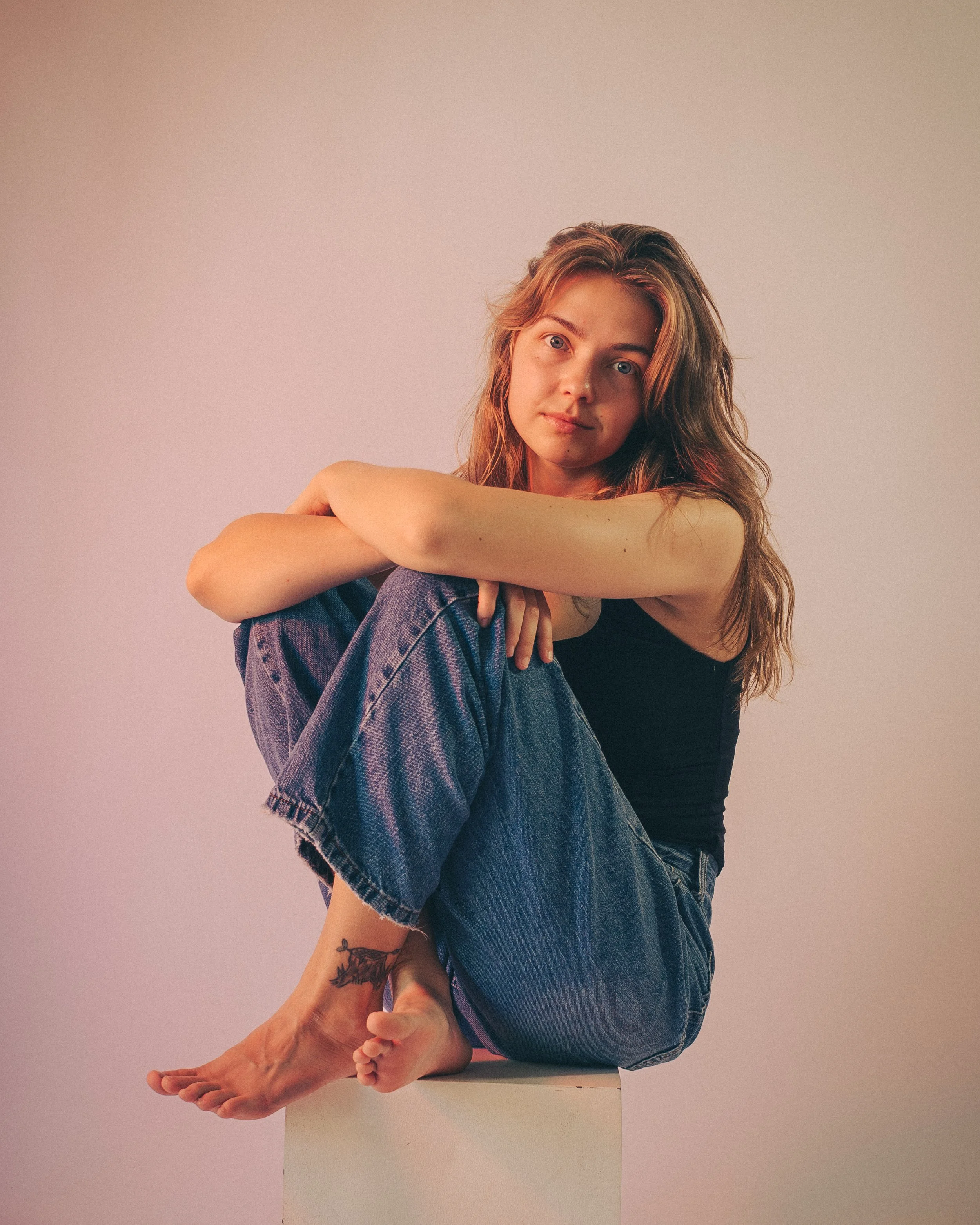 Young woman with long, wavy brown hair, sitting with her knees drawn to her chest, resting her arms on her knees, looking directly at the camera. She is wearing a black sleeveless top and blue jeans, with a tattoo of a leaf on her ankle visible. She 