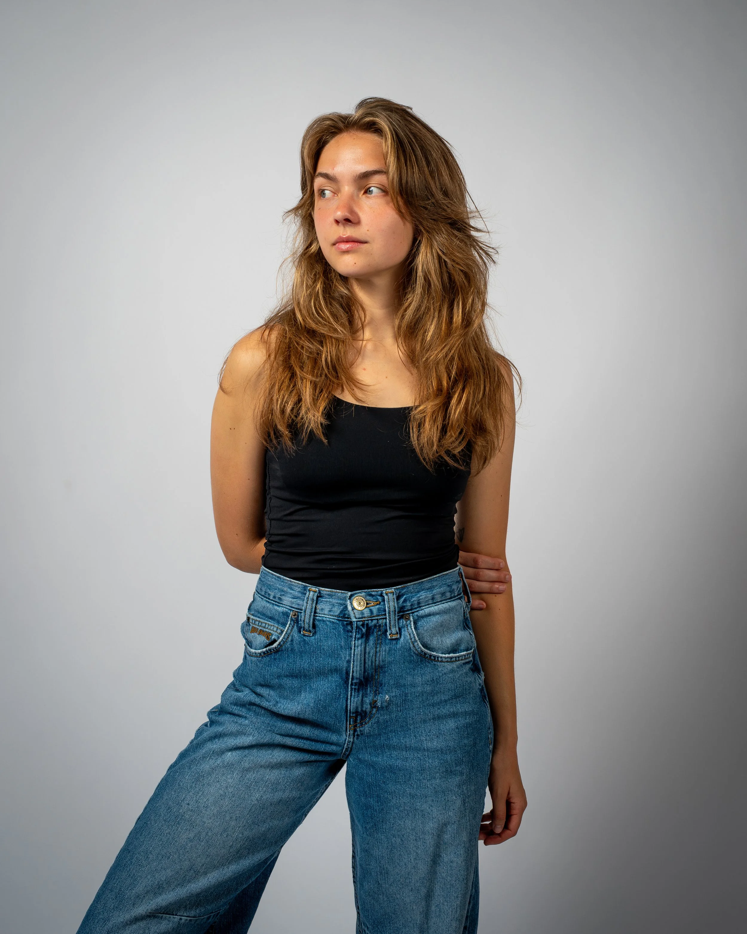 A young woman with long, wavy, light brown hair wearing a black sleeveless top and high-waisted blue jeans standing against a plain gray background.