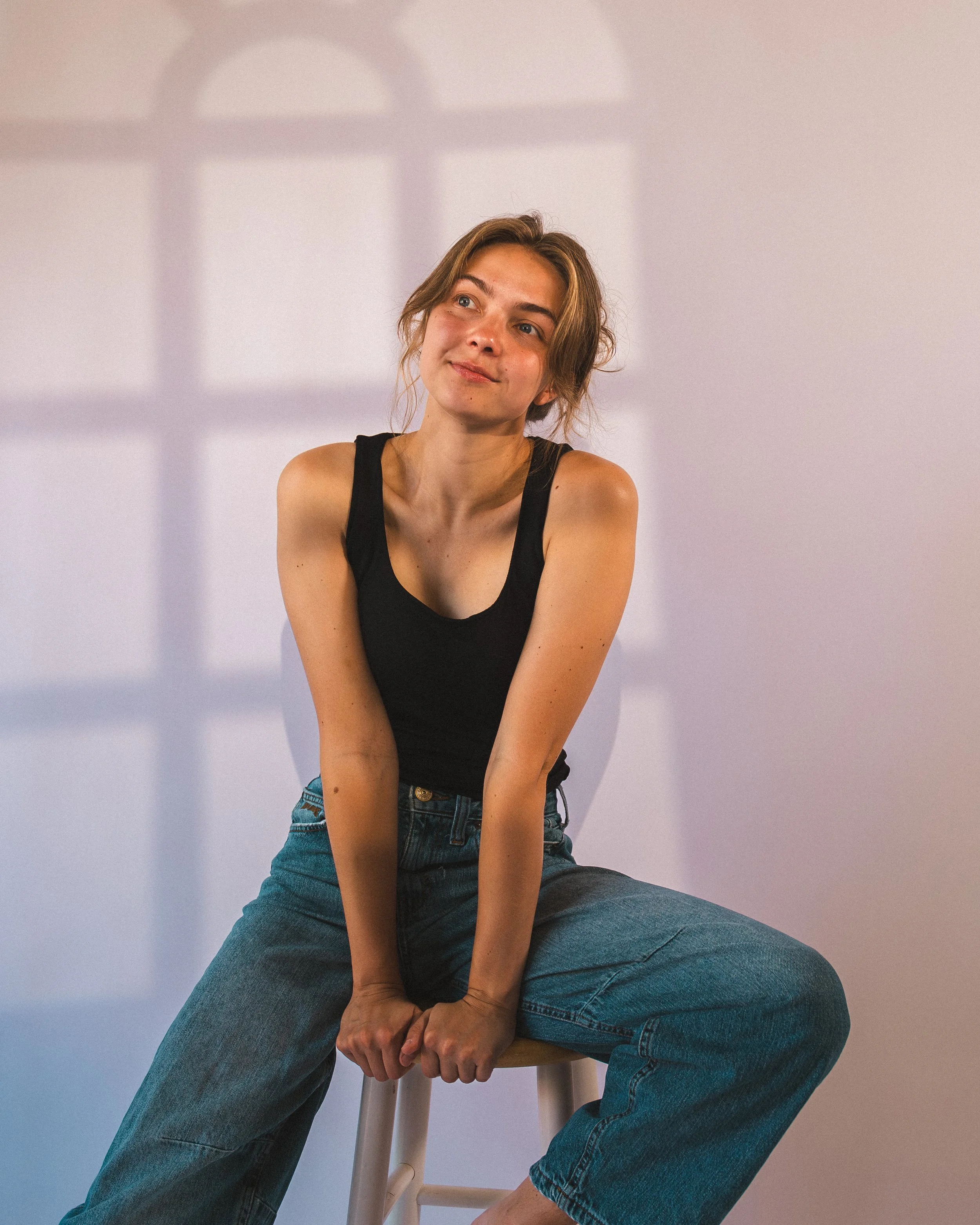 A young woman with short, wavy hair sitting on a wooden stool against a plain light purple background. She is wearing a black tank top and blue jeans, with a relaxed and contemplative expression, looking slightly to her right with her arms resting on