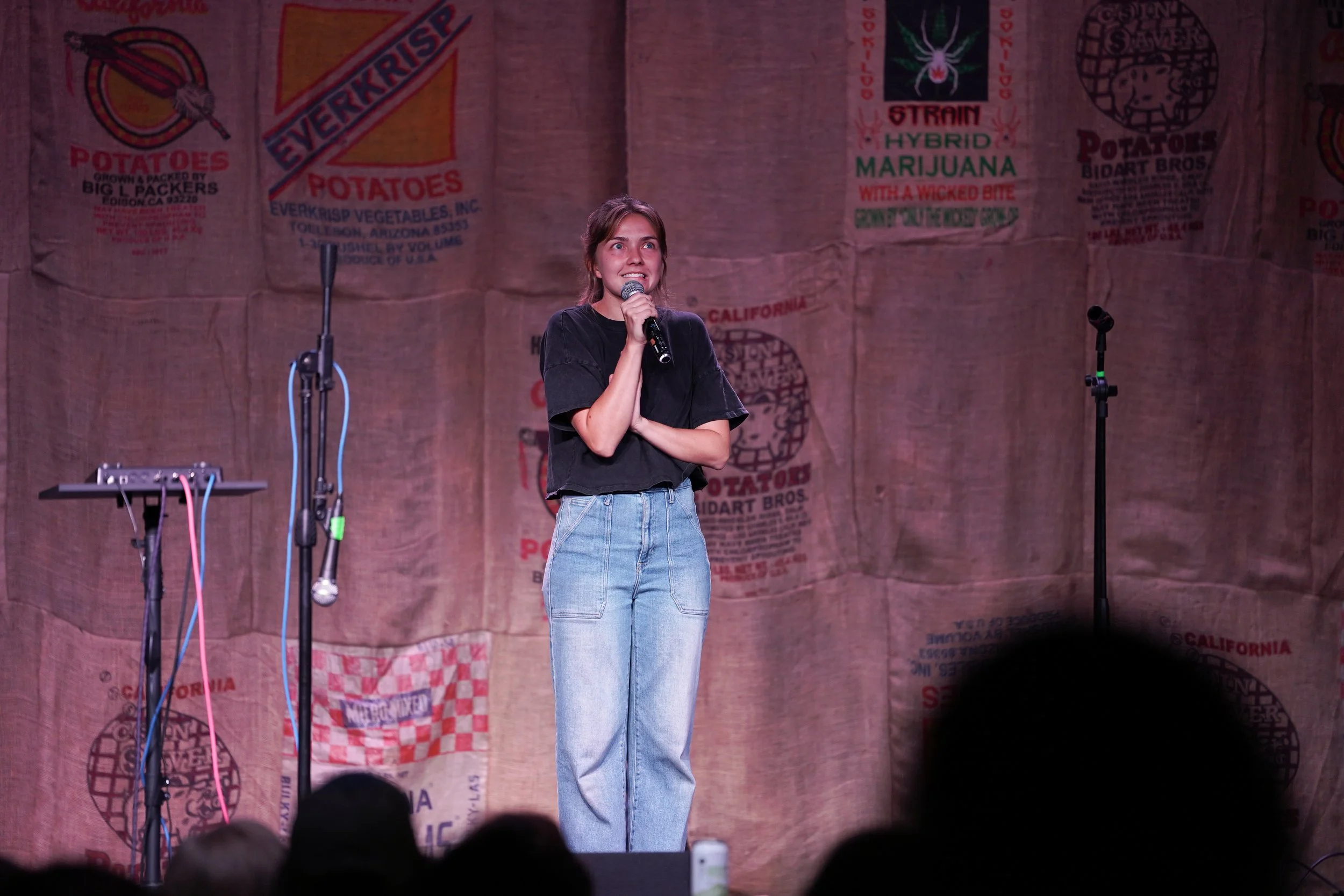 A young woman with brown hair standing on a stage with a microphone in her hand, wearing a black t-shirt and jeans, speaking to an audience. The background features a large burlap banner with various colorful logos and text, including references to p