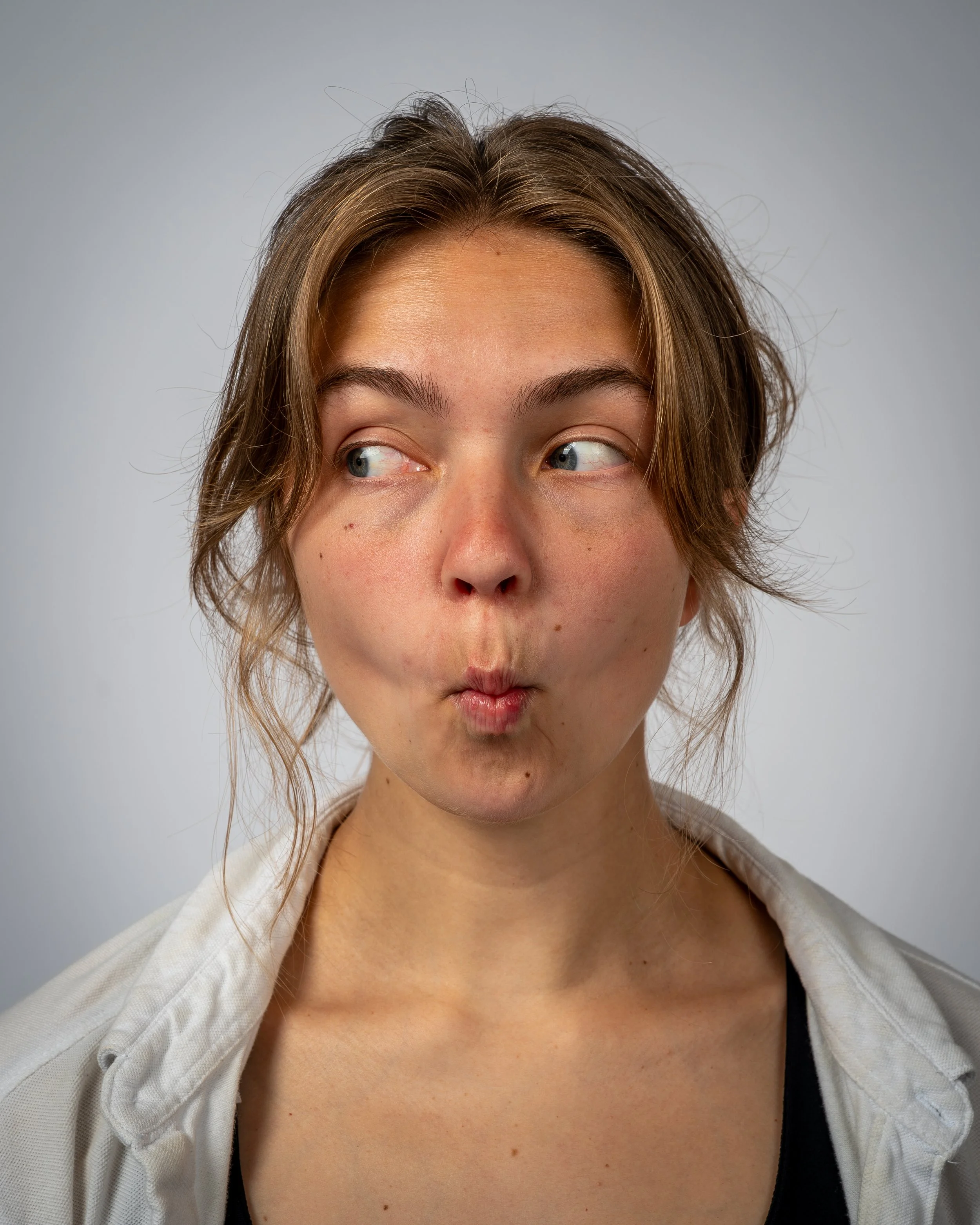 Close-up of a young woman with short, wavy brown hair and blue eyes, making a playful facial expression with pursed lips, wearing a white cardigan over a black top, against a neutral grey background.